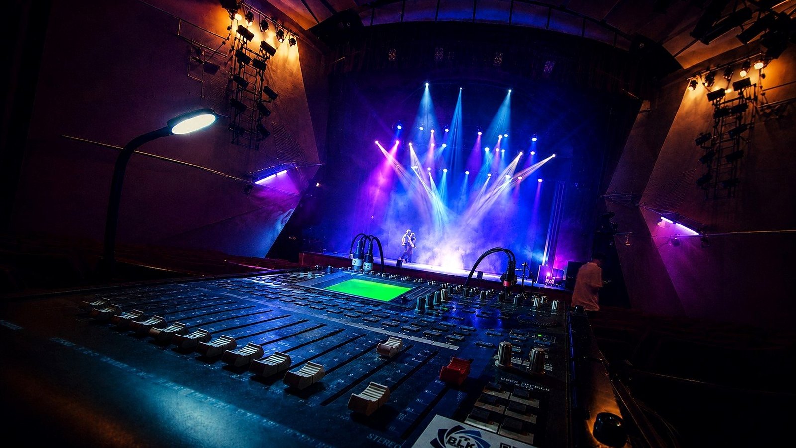 Show lumières et sonorisation pour un gala de danse par BLK Production, console de mixage Yamaha au premier plan avec lampe articulée, scène théâtrale en arrière-plan avec faisceaux bleus et violets, danseurs sur scène, fumée scénique, structures porteuses latérales équipées de projecteurs