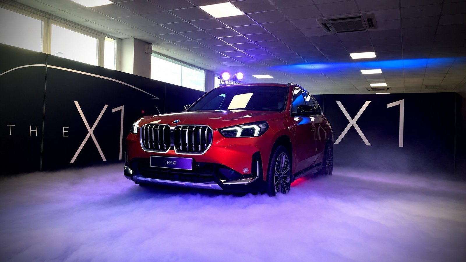 Inauguration BMW X1 rouge par BLK Production en showroom, véhicule sur fond noir avec lettrage 'THE X1' rétro-éclairé, fumée lourde au sol, éclairage scénographique violet et bleu, lyre asservie posée sur le toit du véhicule