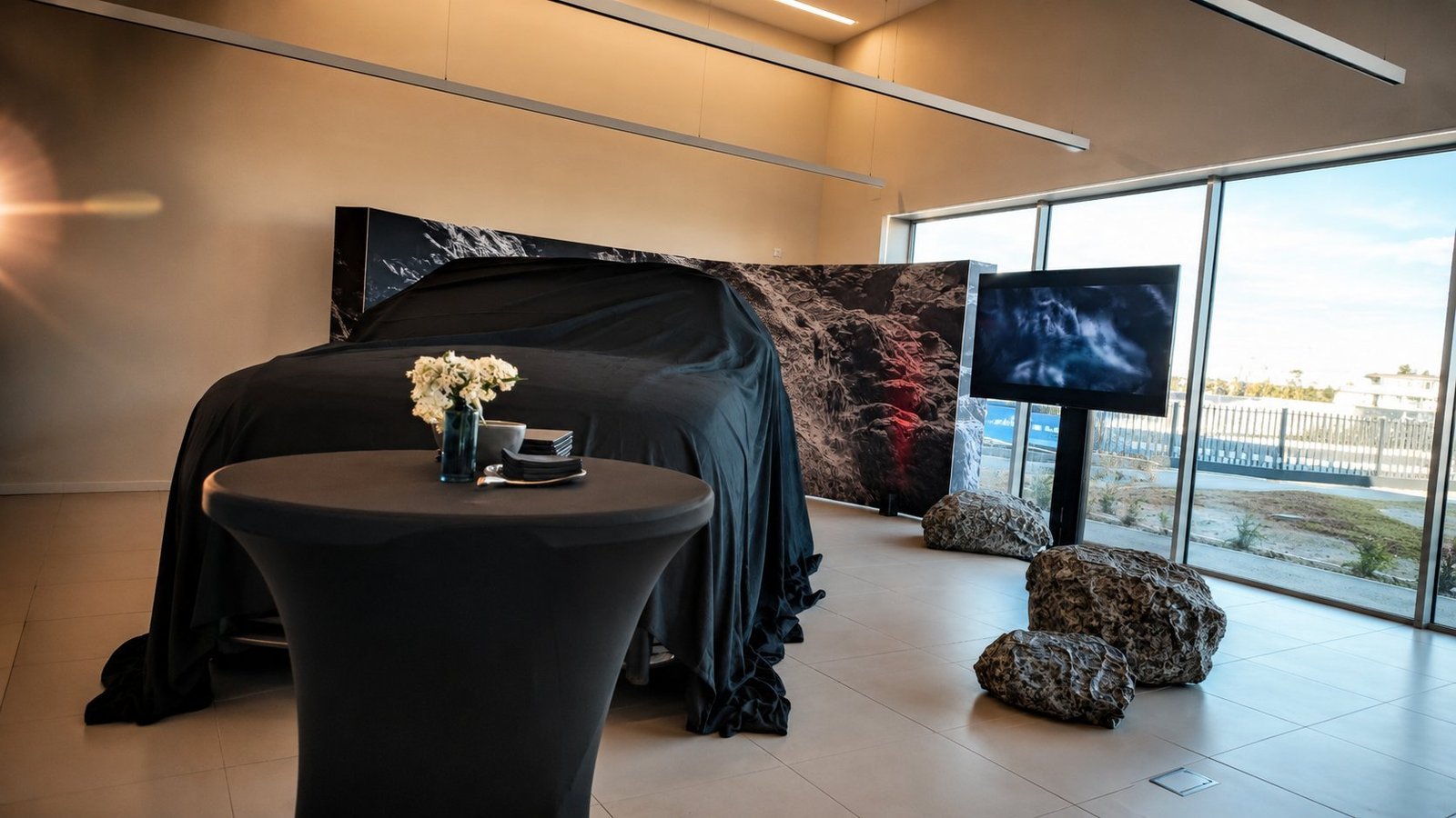 Dévoilement d'un véhicule sous bâche noire par BLK Production lors d'une inauguration showroom, panneau visuel paysage volcanique en arrière-plan, écran TV de présentation et mange-debout habillé en spandex noir
