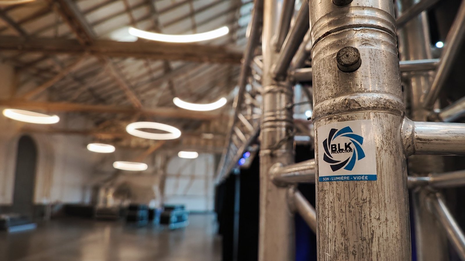 Installation de structure aluminium Global Truss par BLK Production dans une halle industrielle à charpente apparente, gros plan sur un poteau de structure avec sticker logo BLK Production son lumière vidéo, éclairages circulaires LED en arrière-plan