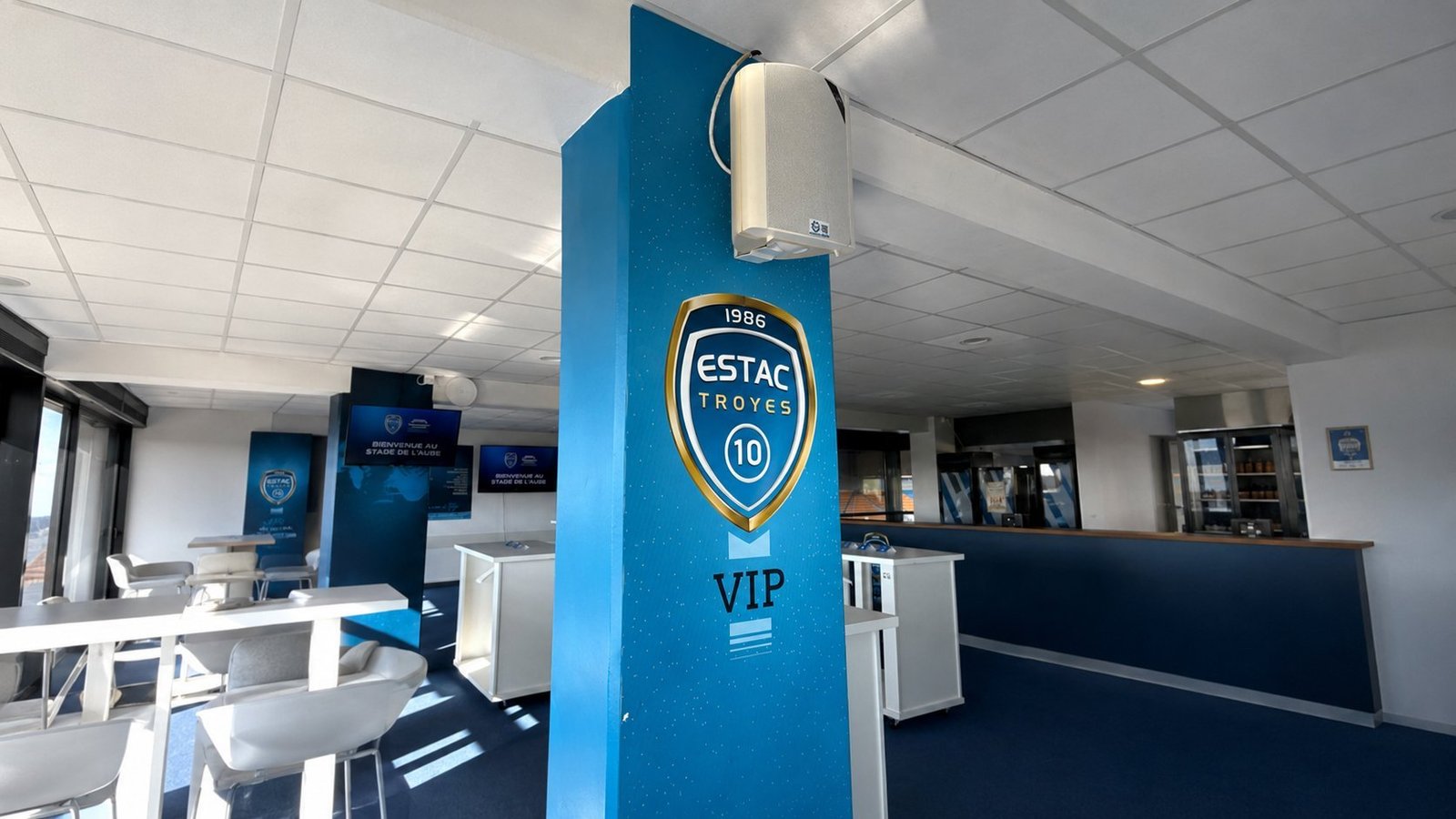 Intégration sonorisation par BLK Production dans les espaces VIP de l'ESTAC Troyes : enceinte de plafond Electro-Voice intégrée sur une colonne d'habillage corporate aux couleurs du club avec écusson ESTAC 1986 et mention VIP, salon haut de gamme avec écrans muraux d'accueil au stade de l'Aube