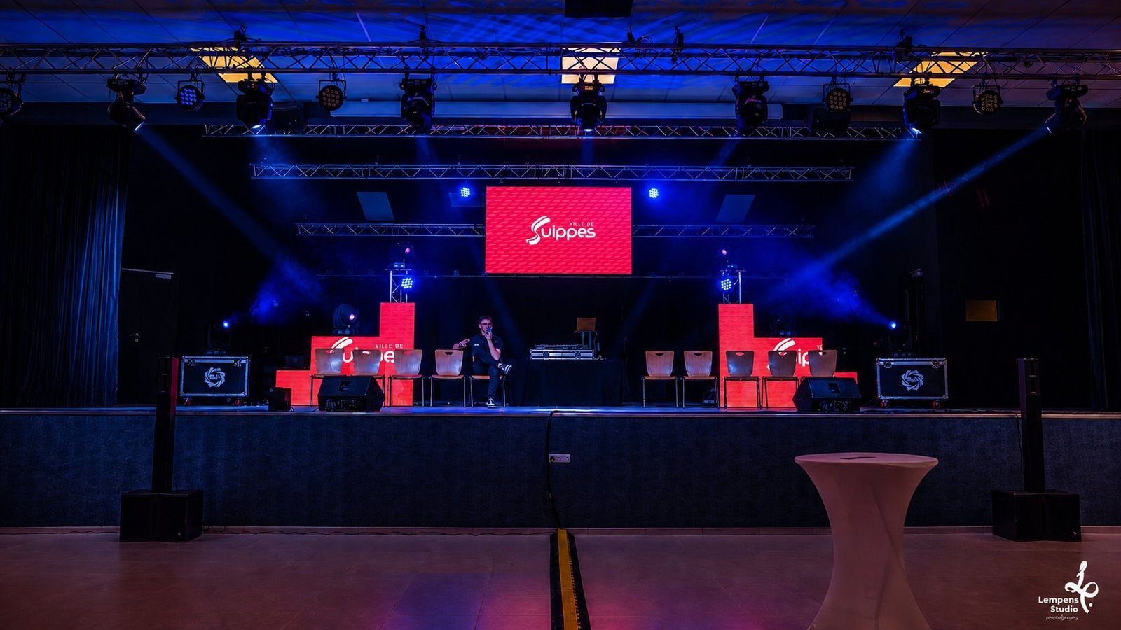 Scénographie lumière complète signée BLK Production pour la Ville de Suippes, lyres asservies bleues en éventail, pars LED, contraste avec écrans LED rouges au logo Ville de Suippes, structure aluminium au plafond, ambiance institutionnelle théâtralisée