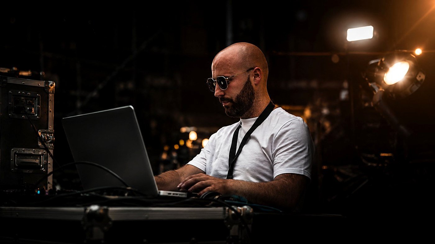 Flavien Lebel en régie technique sur un événement BLK Production, concentré sur ordinateur portable avec lunettes de soleil, lumière de scène en arrière-plan