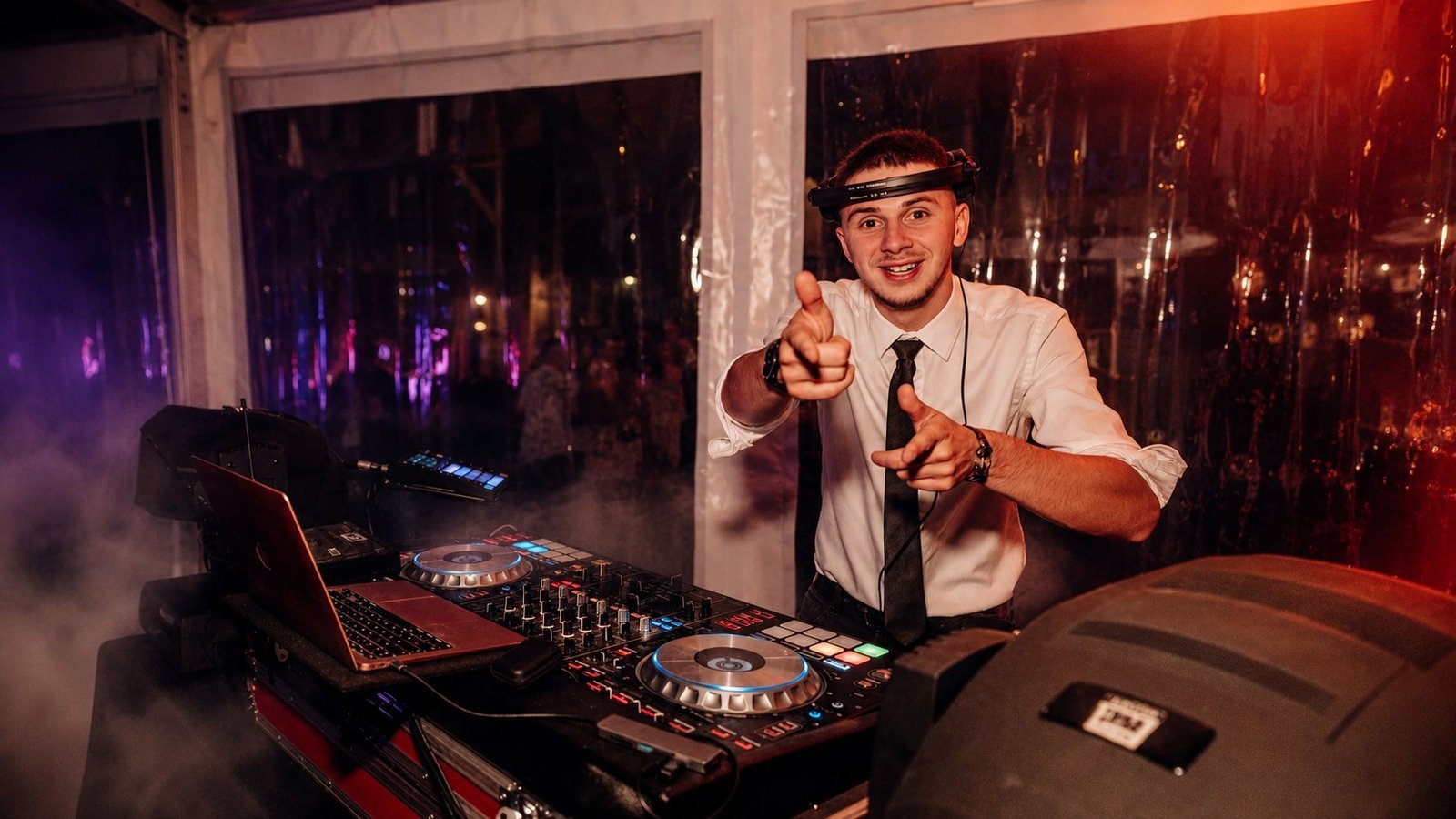 Hugo Verdun en animation DJ sur un événement BLK Production, casque sur la tête, chemise blanche et cravate, derrière sa régie Pioneer avec MacBook, fumée et lumières d'ambiance violettes en arrière-plan